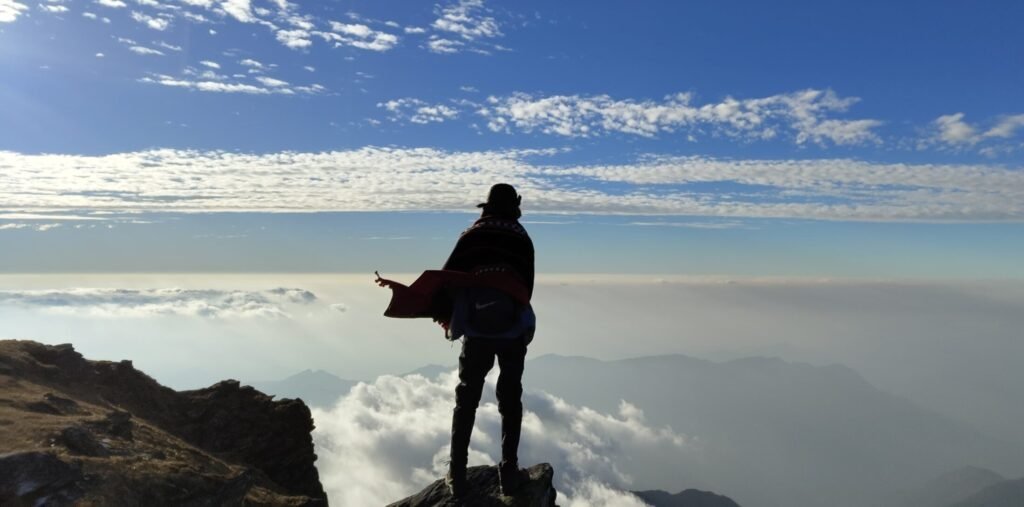 A trekker on a rocky ridge at Mount Saramati, looking over clouds and distant mountain ranges.